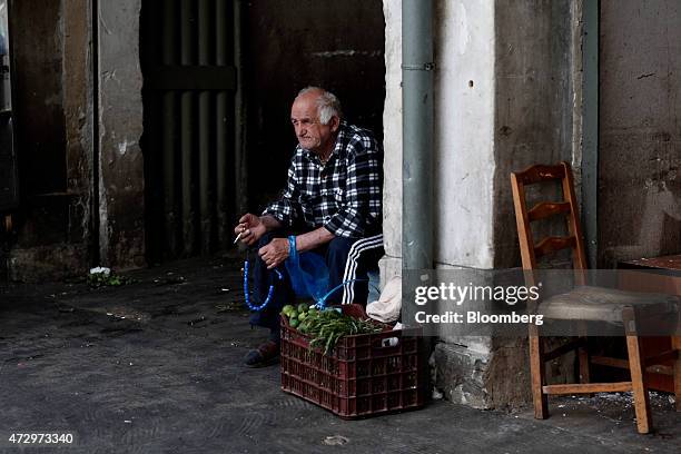 Street vendor smokes a cigarette and holds worry beads as he waits for customers for his box of vegetables at the entrance to Varvakeios fish and...