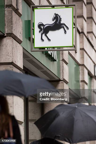 Pedestrians shelter under umbrellas as they pass beneath a Lloyds' prancing horse logo hanging outside a Lloyds Bank branch, a unit of Lloyds Banking...