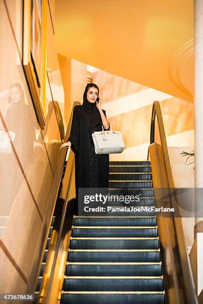 emirati woman talking on cellphone, riding escalator in luxury hotel - gold purse stock pictures, royalty-free photos & images