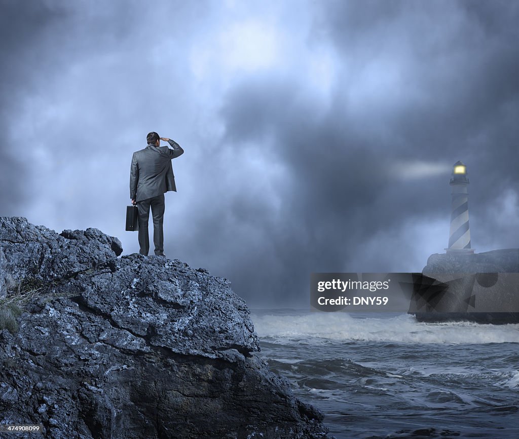 Businessman standing at edge of water looking towards a lighthouse