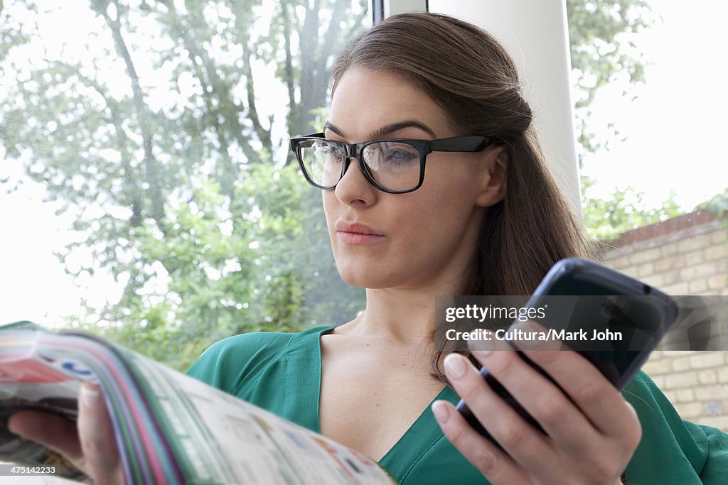 Young woman wearing glasses reading magazine holding cell phone