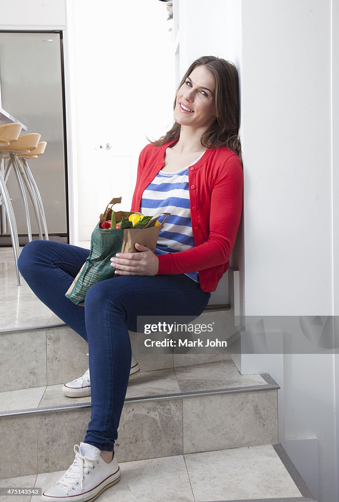 Young woman sitting on kitchen step with grocery bag
