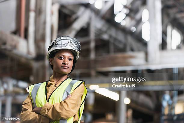 african american woman wearing hardhat and safety vest - factory workers stockfoto's en -beelden