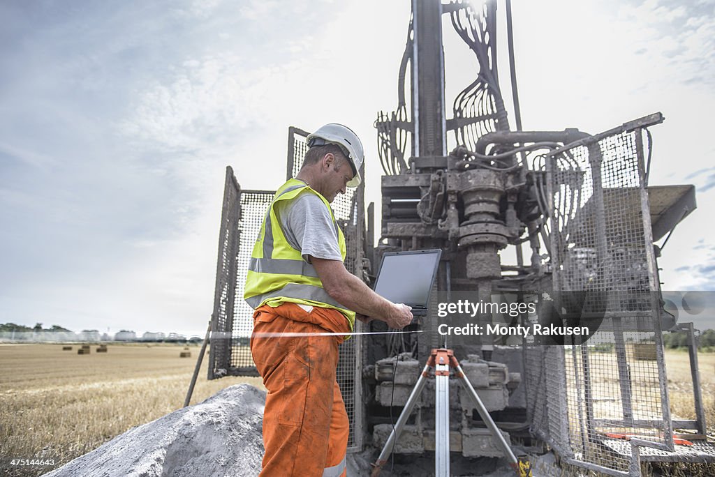 Worker using laptop to survey drilled hole made by drilling rig in field