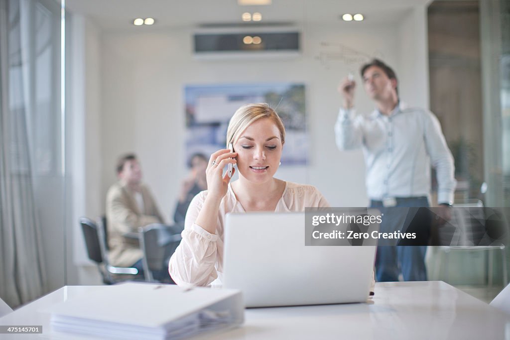 Woman sitting at conference room table using laptop and mobile phone, colleagues working in background