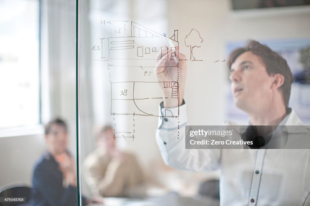 Man drawing architectural plans on glass wall, colleagues in background