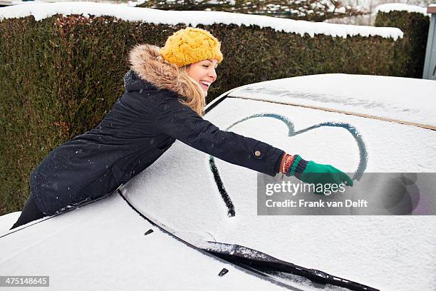 woman drawing heart shape on snow covered windscreen - car window stock pictures, royalty-free photos & images