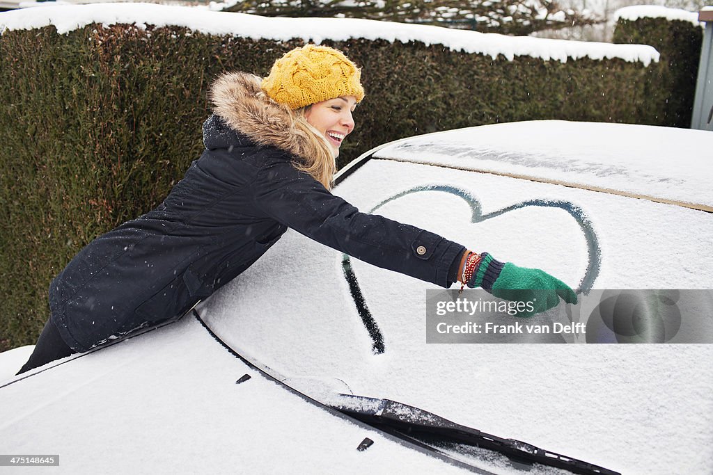 Woman drawing heart shape on snow covered windscreen