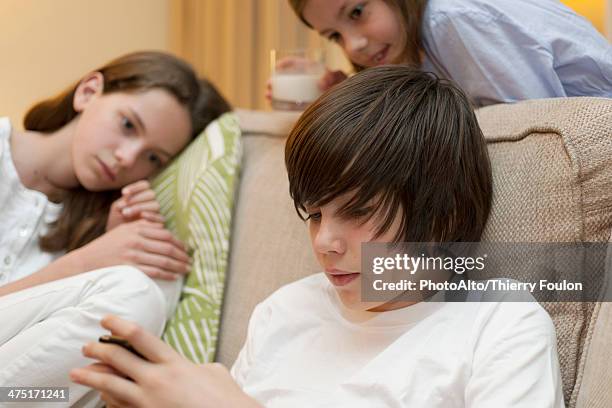 teenage boy using smartphone while sisters watch in background - famiglia multimediale foto e immagini stock