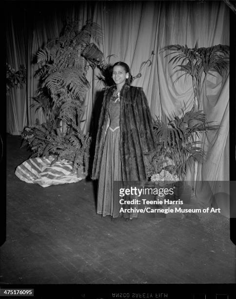 Woman modeling sheer gown and fur coat over shoulders, on stage with potted plants for sixth annual Beauty Shop Owners Fashion Show, Schenley High...