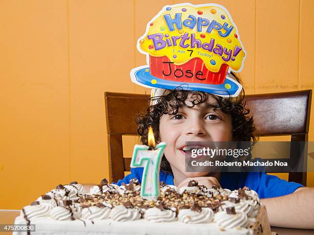 Latino boy with cake, celebrating his seventh birthday, seated on a wooden chair and wearing a birthday cap.