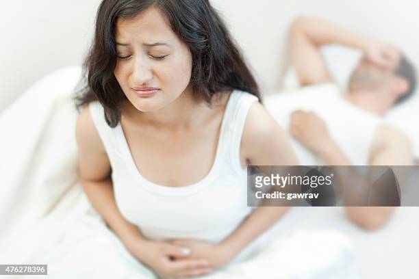 grimacing woman holding her aching stomach and unaware husband sleeping. - pms stock pictures, royalty-free photos & images