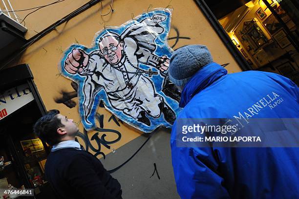 People look at a street art mural showing Pope Francis as a superman, flying through the air with his white papal cloak billowing out behind him and...
