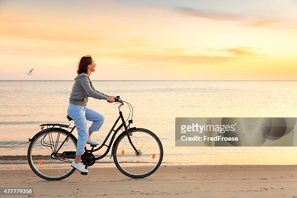 reife frau mit fahrrad am strand bei sonnenuntergang - frau fahrrad stock-fotos und bilder