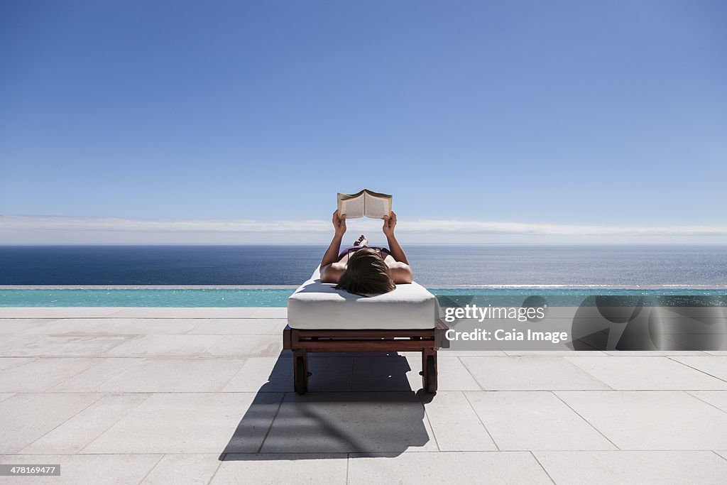 Woman reading on lounge chair at poolside overlooking ocean