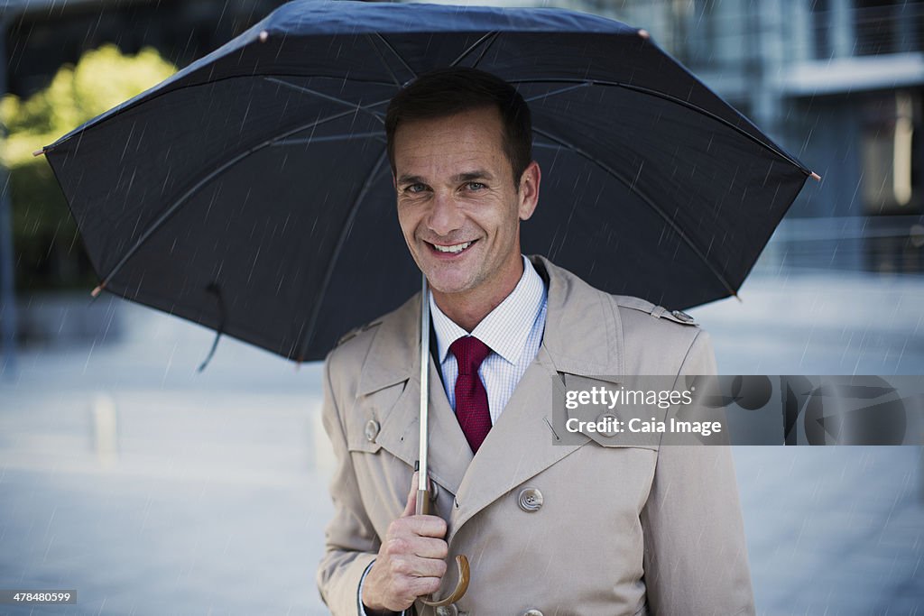 Smiling businessman in trench coat under umbrella