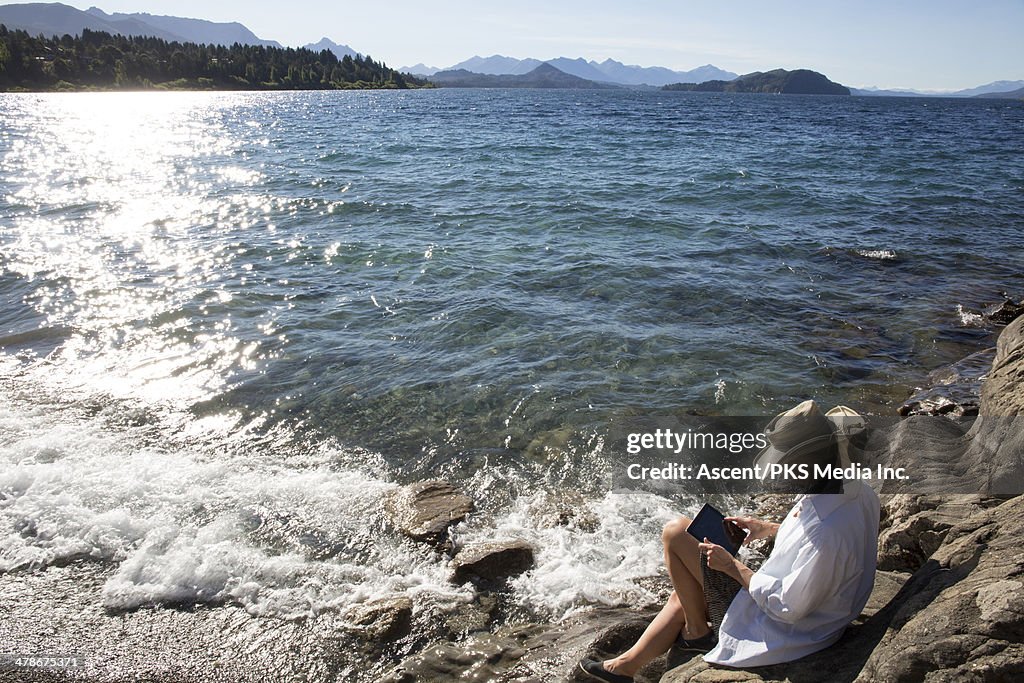 Woman relaxes by lake edge, uses digital tablet