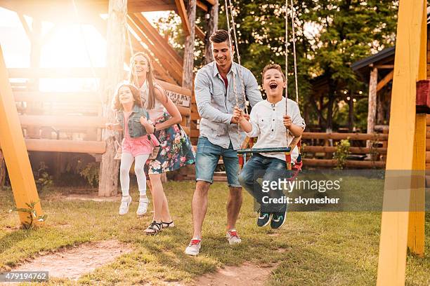 young parents swinging their cheerful children at sunset. - family garden play area stock pictures, royalty-free photos & images