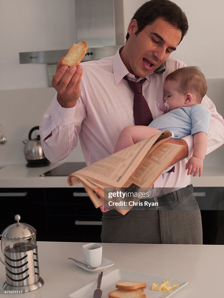 Father talking on phone with toast while holding baby daughter in a kitchen