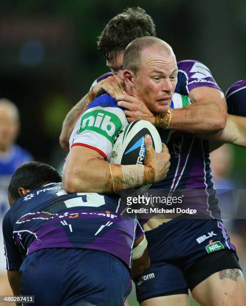 Beau Scott of the Knights is tackled by Will Chambers and Ben Hampton of the Storm during the round three NRL match between the Melbourne Storm and...