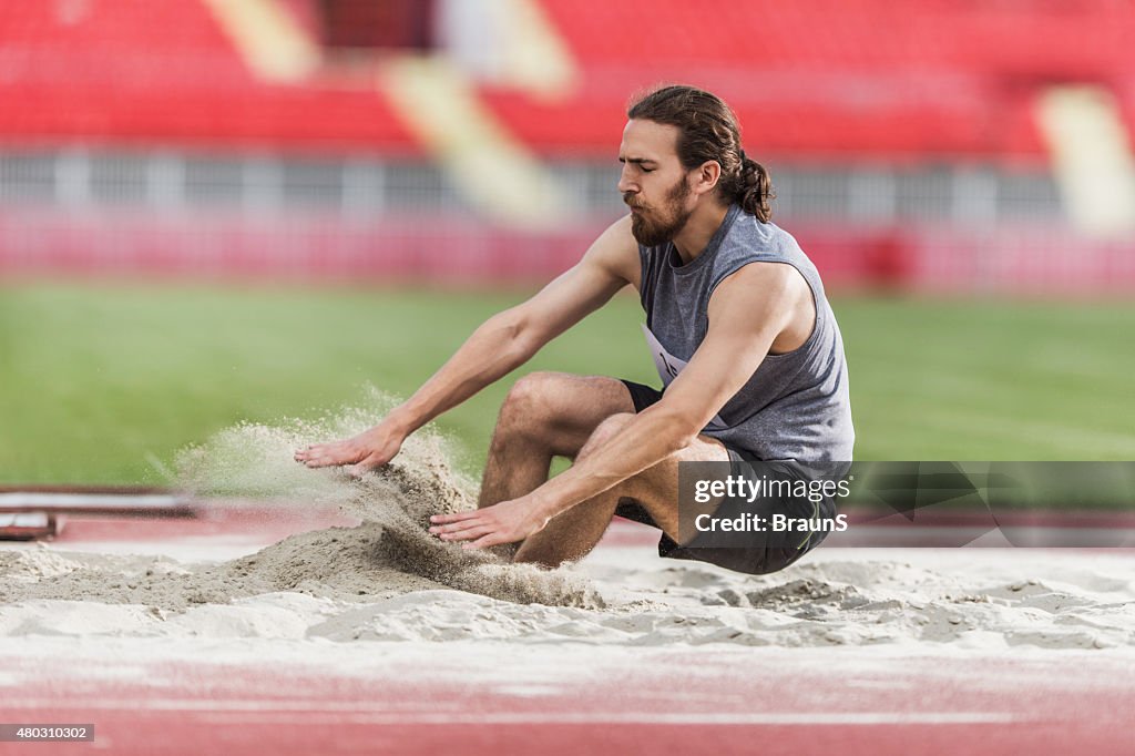 Young man exercising long jumps and landing in a sand.