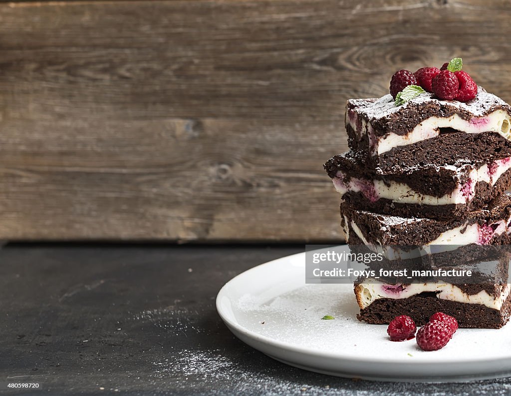 Brownies-cheesecake tower with raspberries on white plate, wooden backdrop