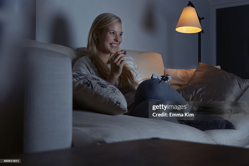 Young woman sitting on sofa, watching tv, eating chocolate