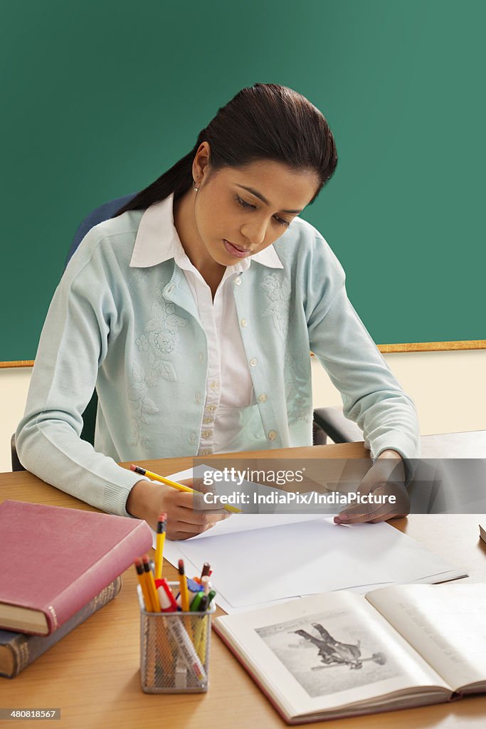 Young female teacher writing on paper at desk in classroom
