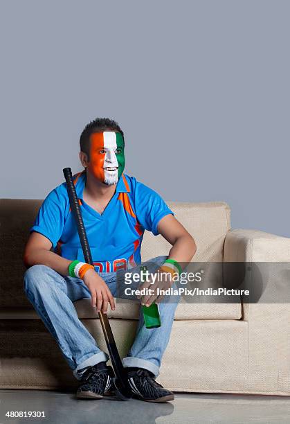 young man with painted face in indian tricolor sitting on sofa with hockey stick and beer bottle - blank sports jersey stock pictures, royalty-free photos & images
