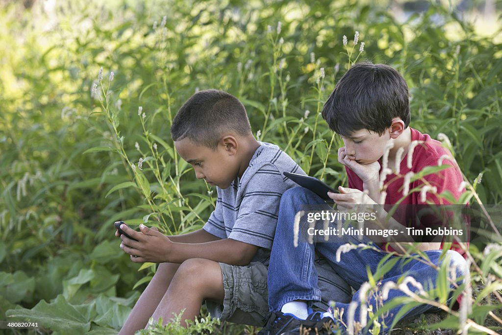 Two boys sitting in a field, one on a smart phone and one using a digital tablet.