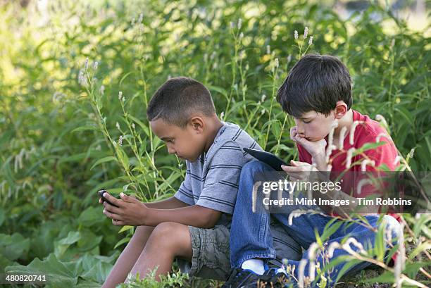 Two Boys Sitting In A Field One On A Smart Phone And One Using A Digital Tablet , Photo