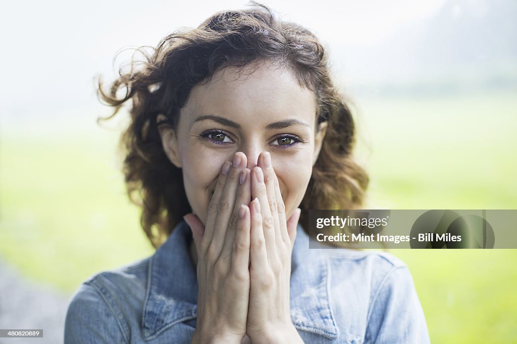 A young woman in a rural landscape, with windblown curly hair. Covering her face with her hands, and laughing.