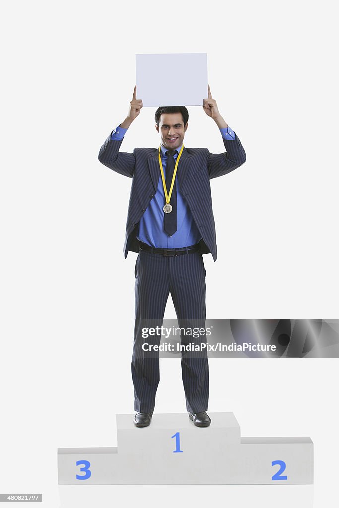 Portrait of confident businessman with medal holding placard while standing on winners podium over white background