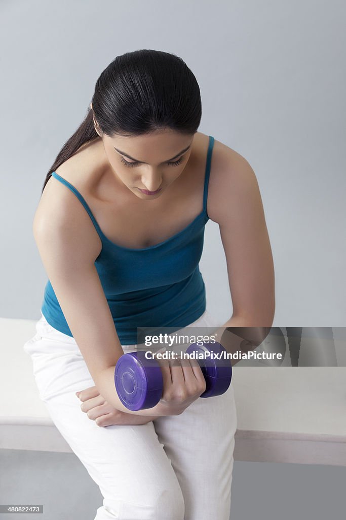 Young woman lifting dumbbells while sitting on bench against gray background