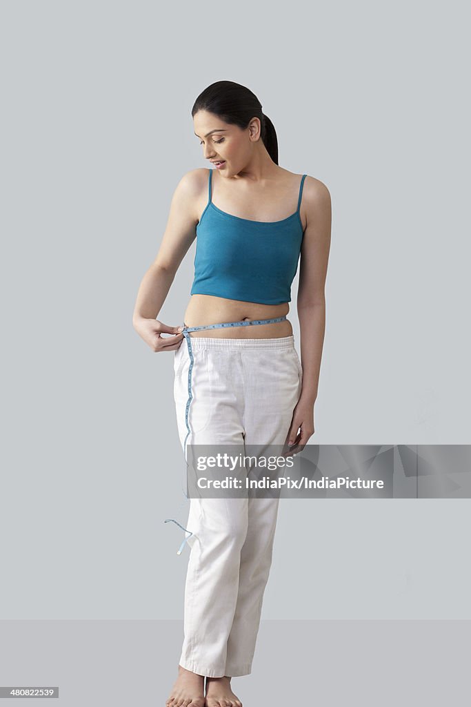 Full length of young woman measuring waist with tape over gray background