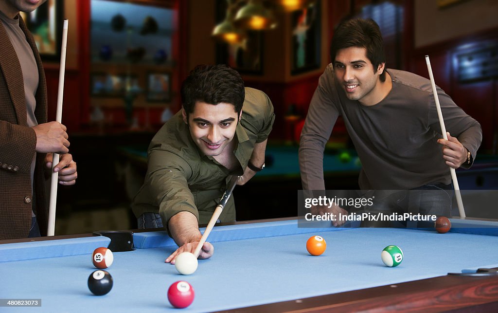 Young man aiming at cue ball while friends standing by pool table in club