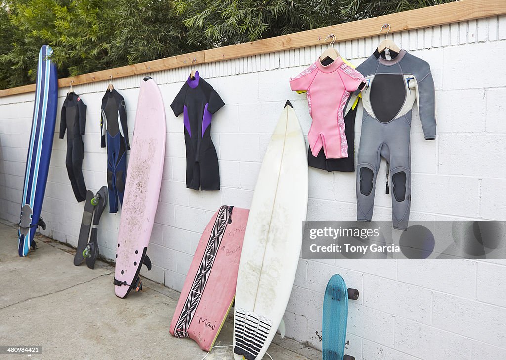 Surfboards and skateboards leaning against wall, and wetsuits hanging