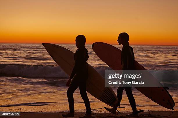 two girls walking along beach at sunset carrying surfboards - redondo beach stock pictures, royalty-free photos & images