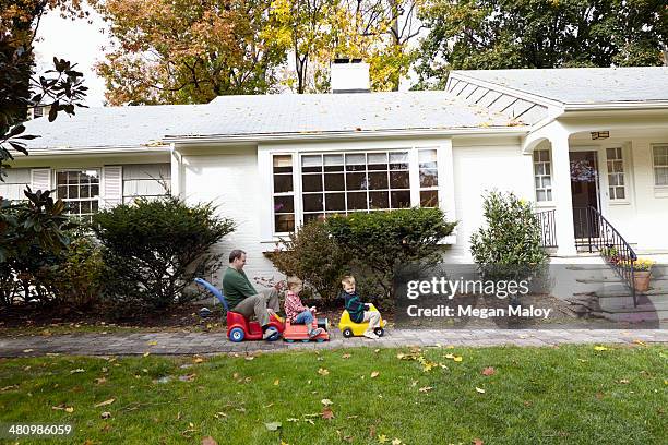 father and young sons riding on toy cars in garden - two kids playing with hose stock-fotos und bilder