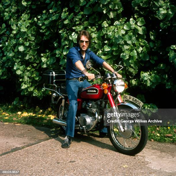 Portrait of British radio disc jockey and broadcaster Johnnie Walker as he poses on a motorcycle, London, England, 1977.