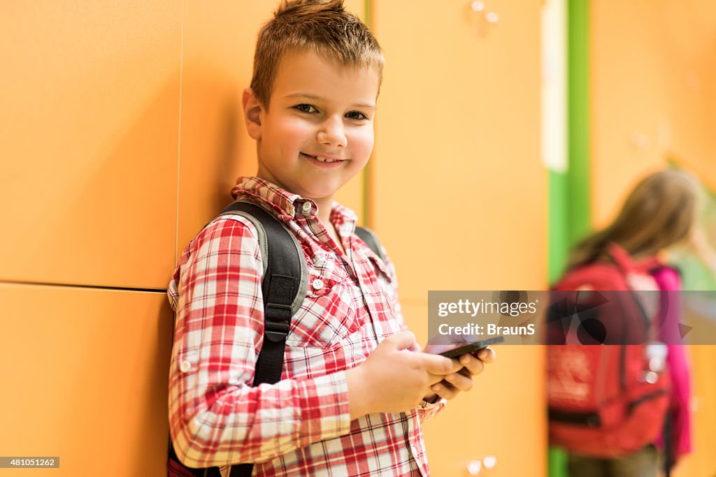 Smiling schoolboy using mobile phone in a school hallway.
