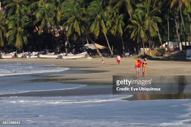 Mexico, Oaxaca, Puerto Escondido, Playa Marinera with couple walking along shore line of boats pulled up onto sand and overhanging palms beyond.