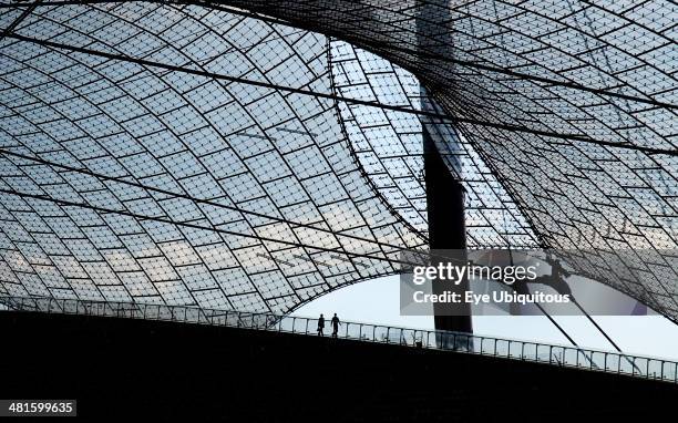 Germany, Bavaria, Munich, Olympic Stadium built as the main venue for the 1972 Summer Olympics. Pair of tourists silhouetted on balcony beneath large...