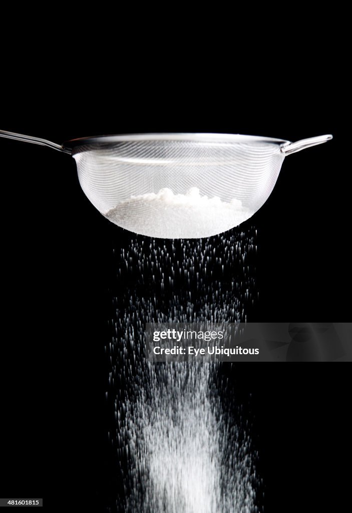 White plain flour being sifted through a sieve against a black background