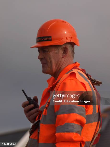 Mature construction worker with walkie talkie directing crane operator, UK.