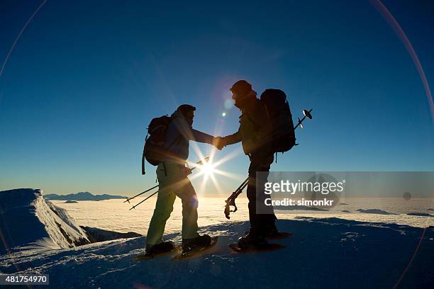 zwei wanderer schütteln hände auf schnee bedeckt landschaft - schneeschuh stock-fotos und bilder