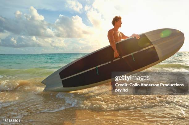 caucasian man carrying paddle board in ocean surf - paddle board men imagens e fotografias de stock