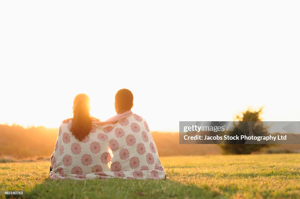 Sun setting behind couple wrapped in blanket in field