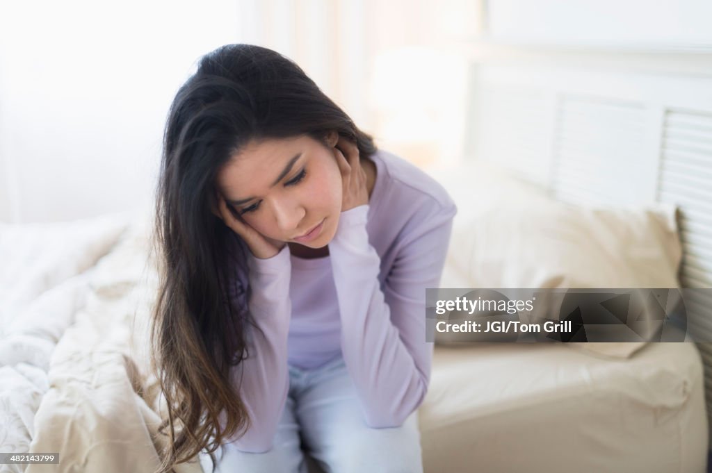 Sad Hispanic woman sitting at edge of bed