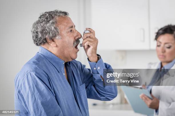 senior man using asthma inhaler in doctor's office - asthmainhalator stock-fotos und bilder